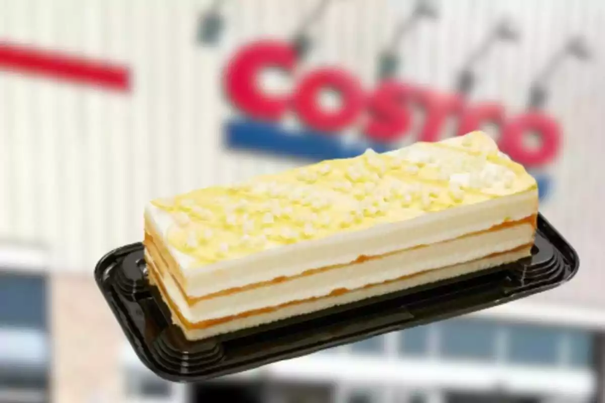 Pastel rectangular de tres capas con betún amarillo y decoraciones blancas sobre una charola negra frente a una tienda Costco desenfocada Pastel rectangular de tres capas con betún amarillo y decoraciones blancas sobre una charola negra frente a una tienda Costco desenfocada