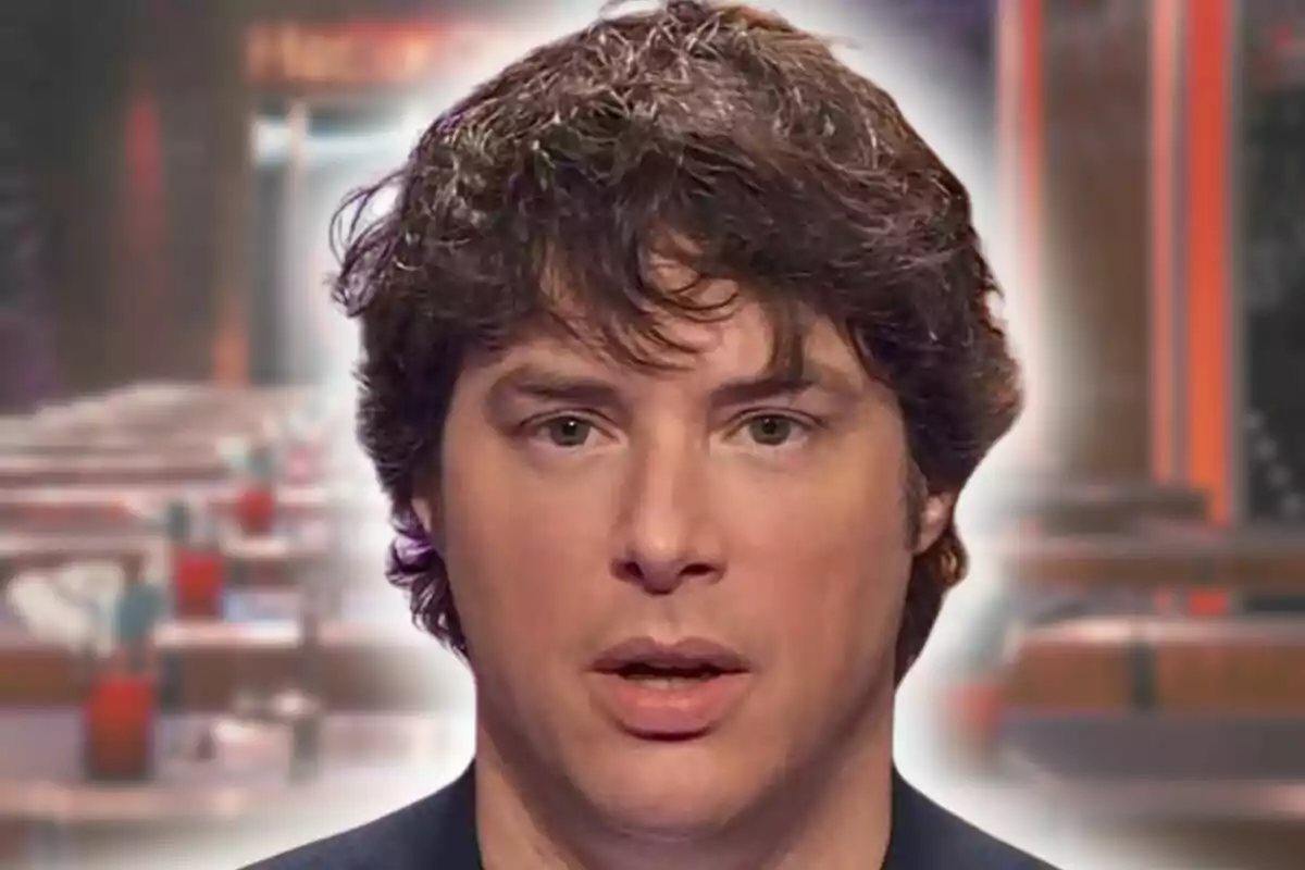 Jordi Cruz con el cabello oscuro y rizado con expresión seria aparece en primer plano con un fondo desenfocado que parece ser un restaurante o cafetería.