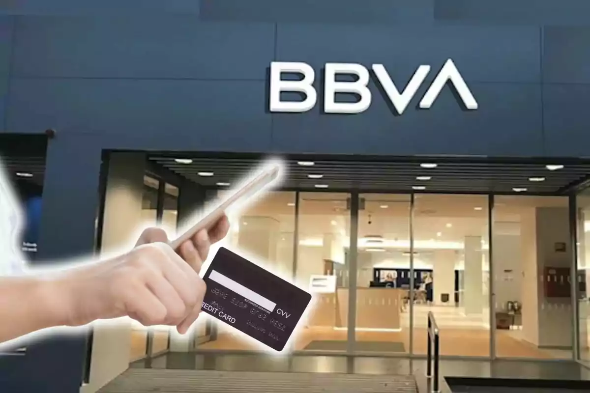 Una persona sostiene un teléfono móvil y una tarjeta de crédito frente a una sucursal del banco BBVA.