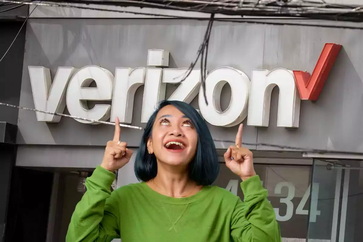 Una persona con cabello azul y suéter verde sonríe y señala hacia arriba frente a un letrero de Verizon.