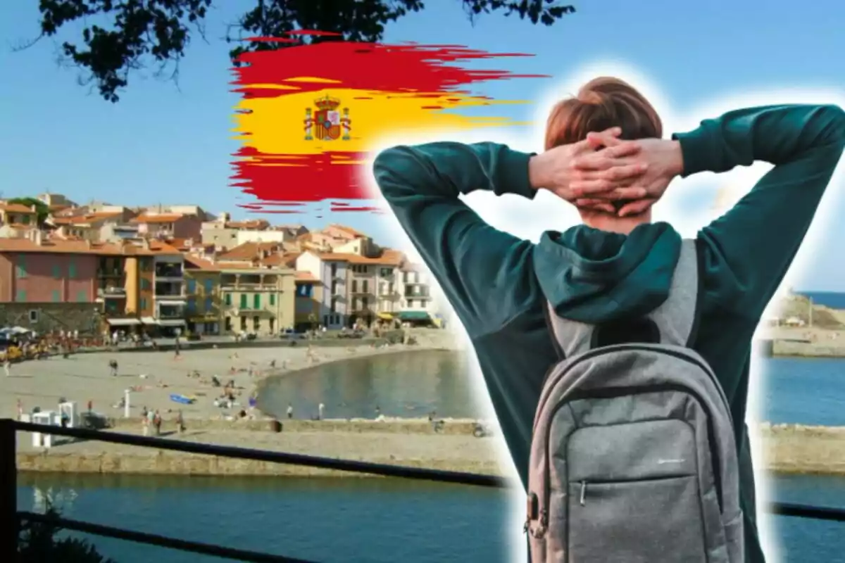 Persona con mochila observando una playa y un pueblo costero con la bandera de España en el cielo.