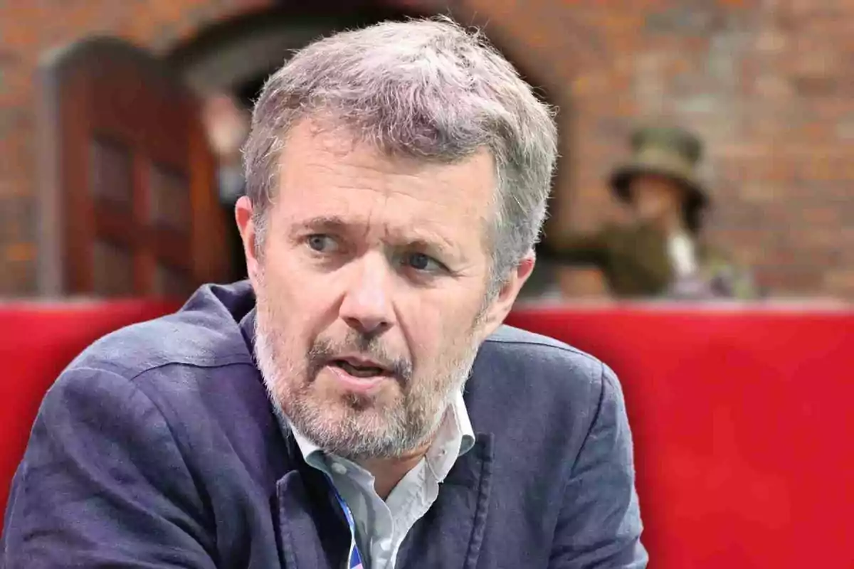 Federico de Dinamarca con cabello canoso y barba corta con expresión seria sentado frente a un fondo desenfocado de ladrillos y una persona al fondo