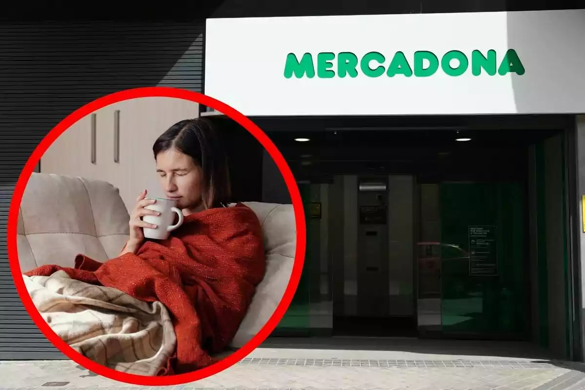 Una persona envuelta en una manta roja sosteniendo una taza mientras está sentada en un sofá con la entrada de una tienda Mercadona al fondo.