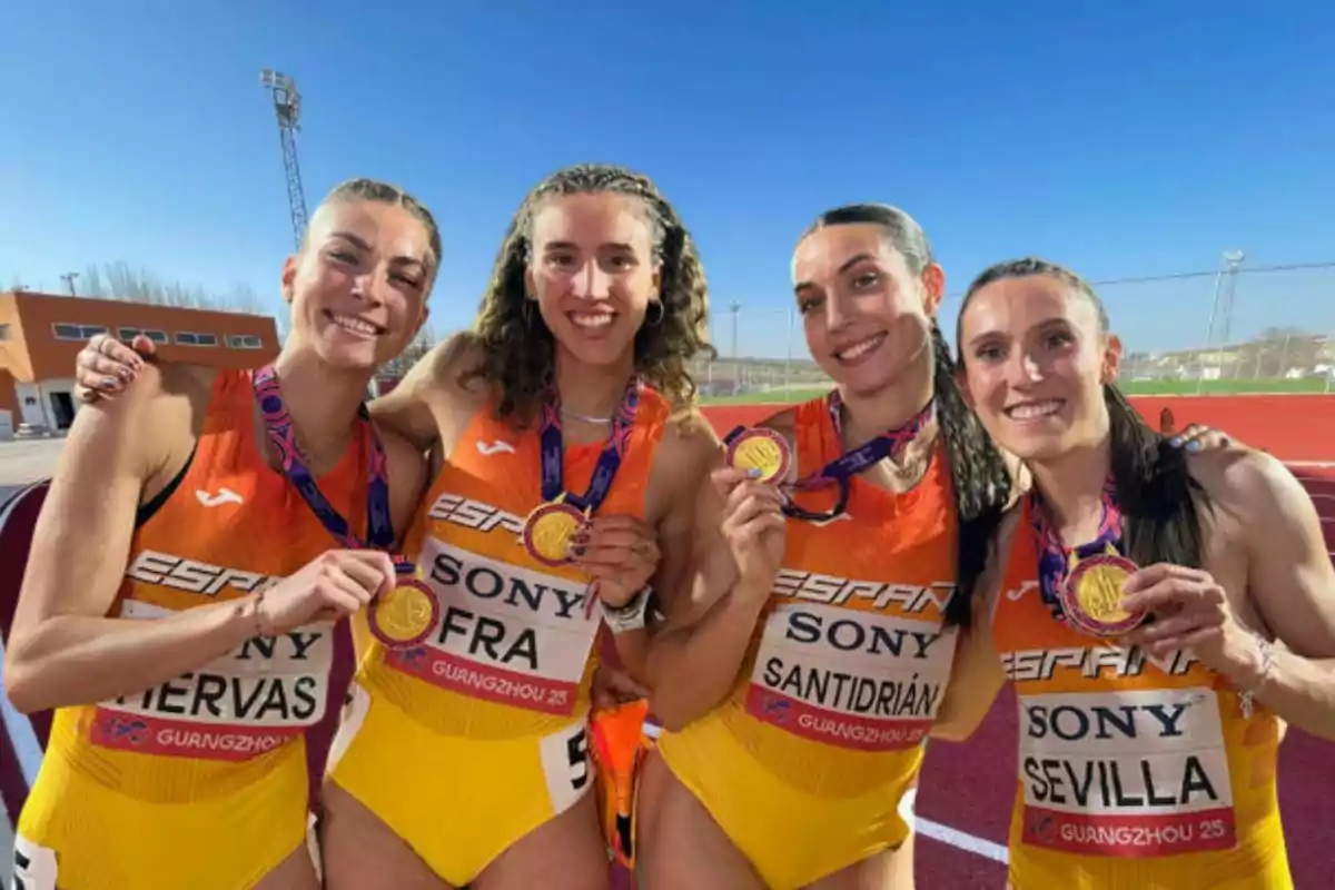 Cuatro atletas sonrientes posan con medallas de oro en una pista de atletismo, vistiendo uniformes naranjas y amarillos.