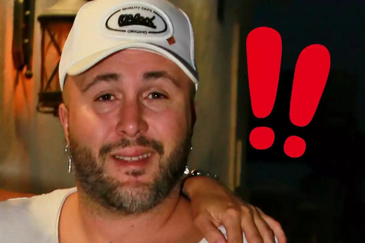 Kiko Rivera con gorra blanca y barba sonríe mientras una mano descansa en su hombro; a su lado hay dos signos de exclamación rojos.