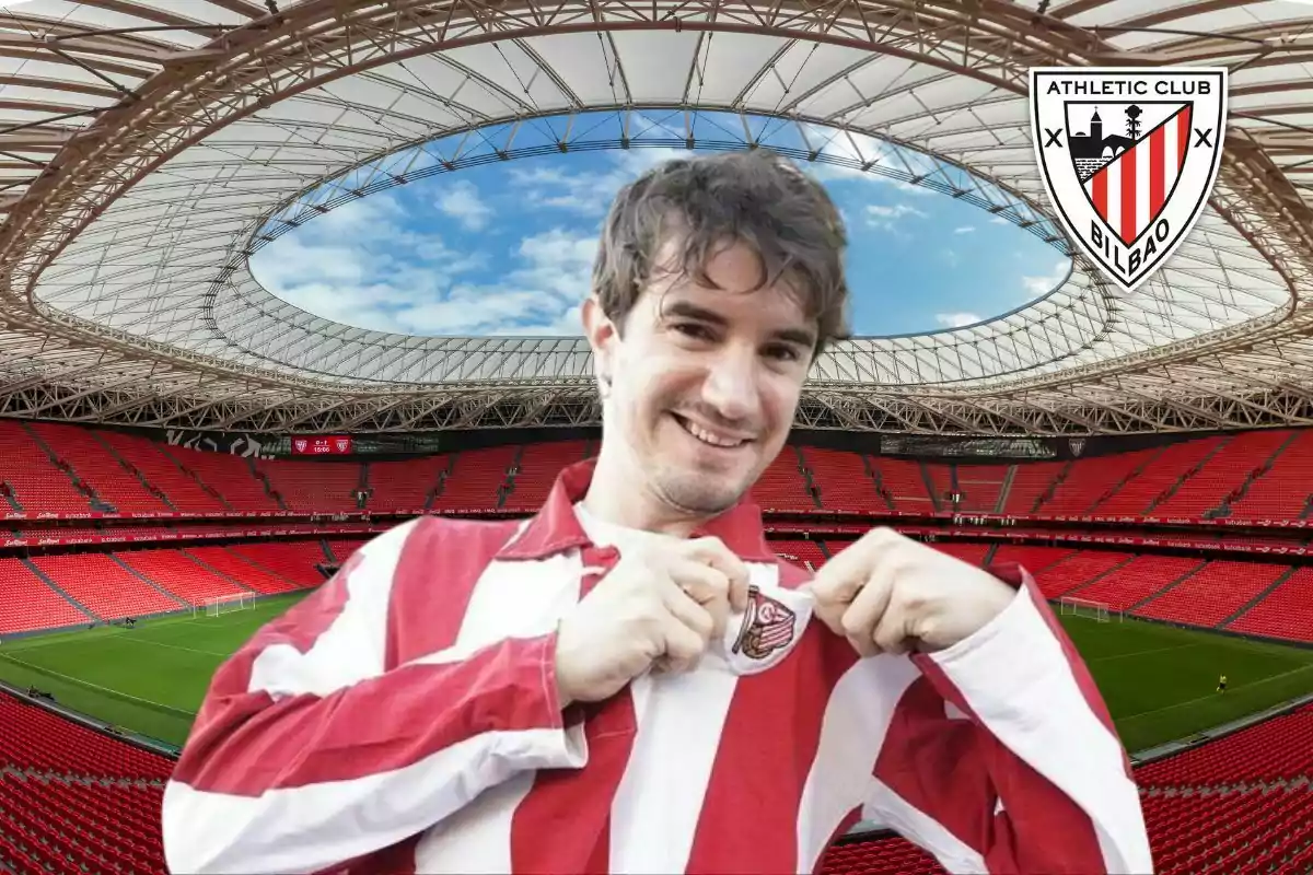 Un hombre sonriente con una camiseta de rayas rojas y blancas sostiene el escudo del Athletic Club de Bilbao en un estadio vacío.