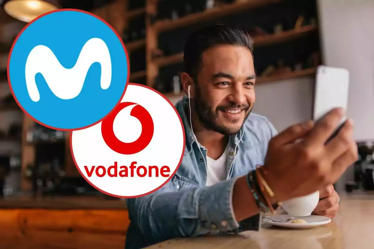 Un hombre sonriente con auriculares sostiene un teléfono móvil mientras está sentado en una cafetería, con los logotipos de Movistar y Vodafone superpuestos en la imagen.