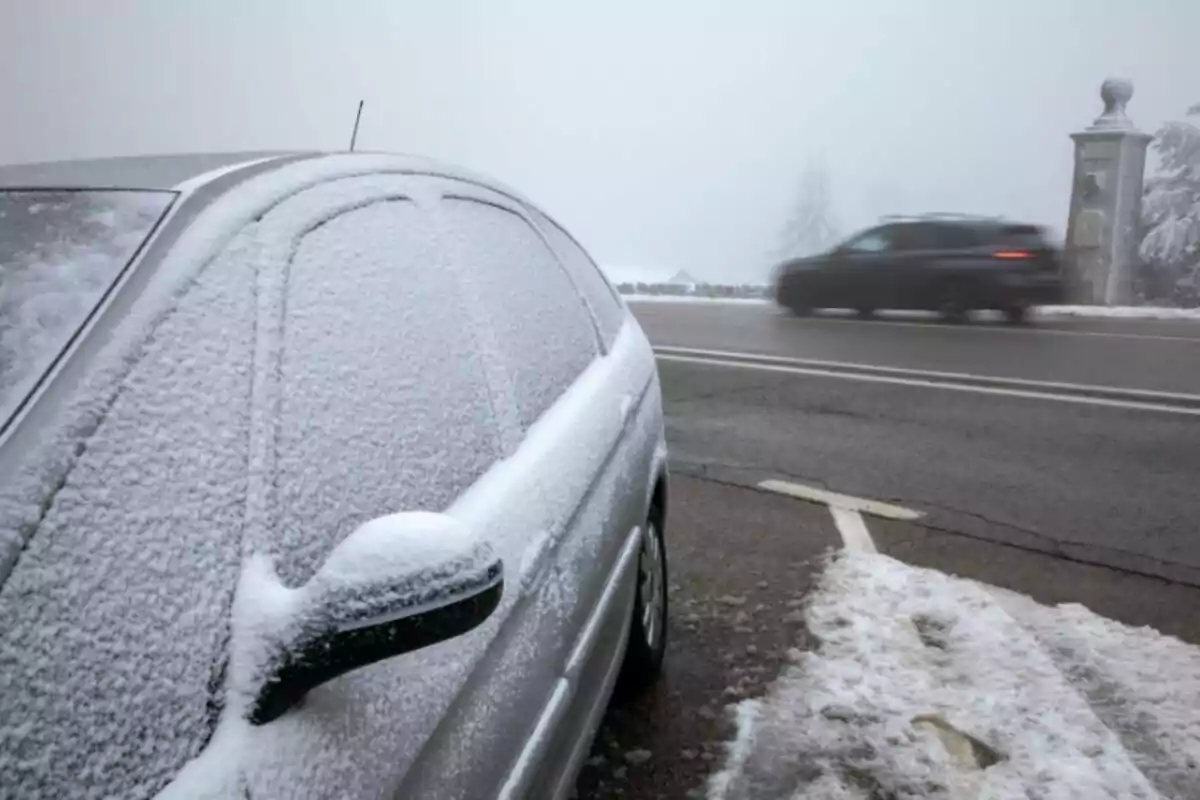 Un coche cubierto de nieve estacionado al lado de una carretera mientras otro vehículo pasa rápidamente en un día nublado.