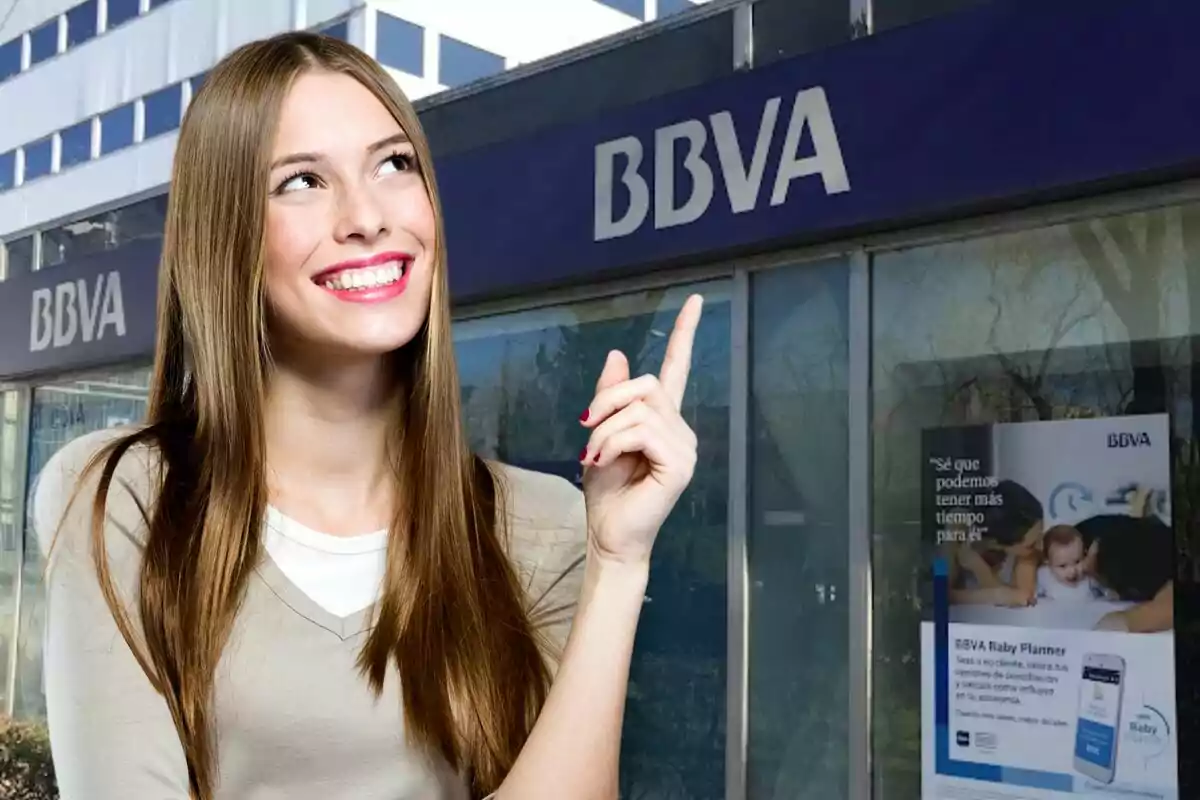 Una mujer sonriente con cabello largo y lacio señala con su dedo índice hacia el letrero de una sucursal de BBVA.
