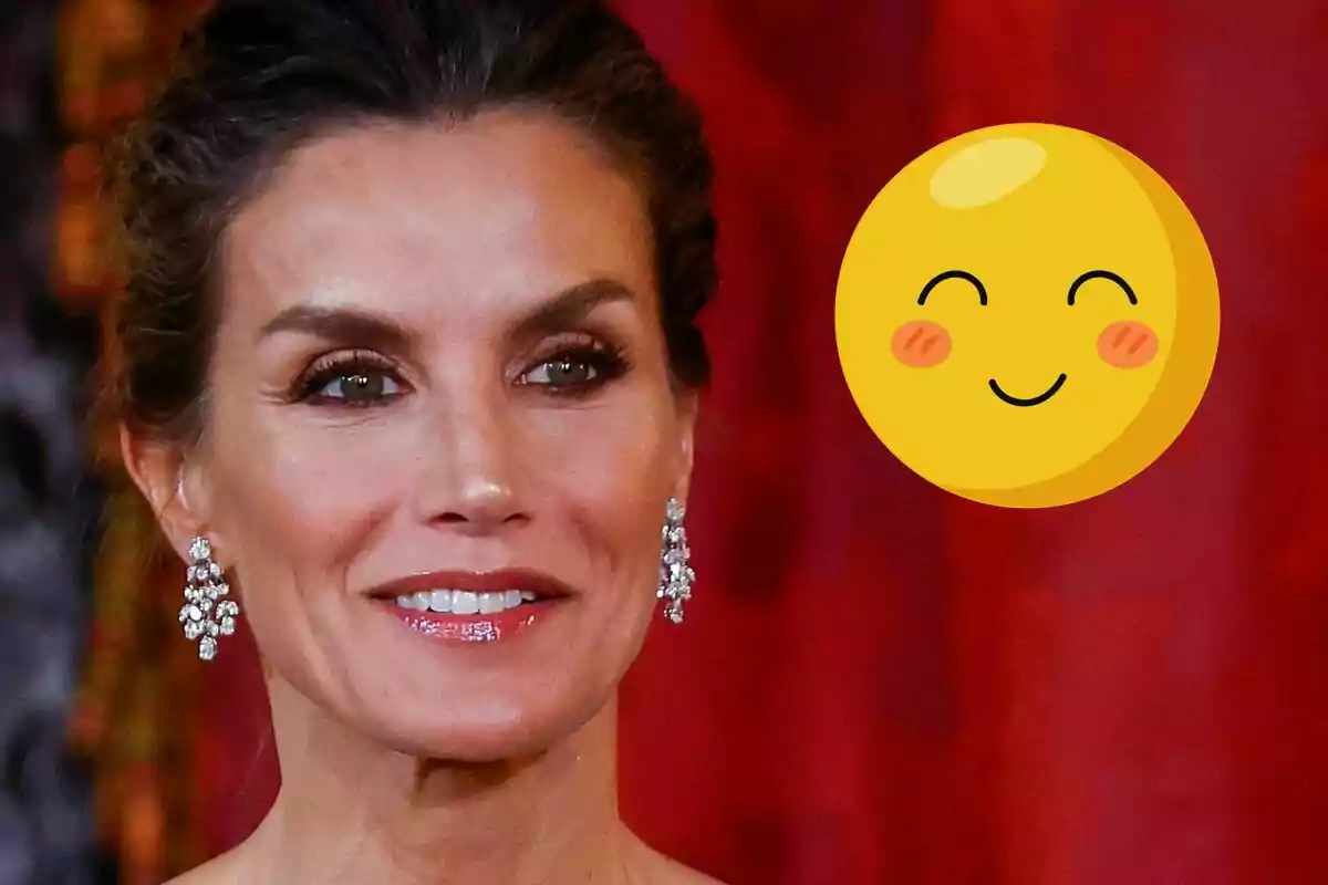 La reina Letizia con aretes brillantes sonríe frente a un fondo rojo, acompañada de un emoji sonriente.
