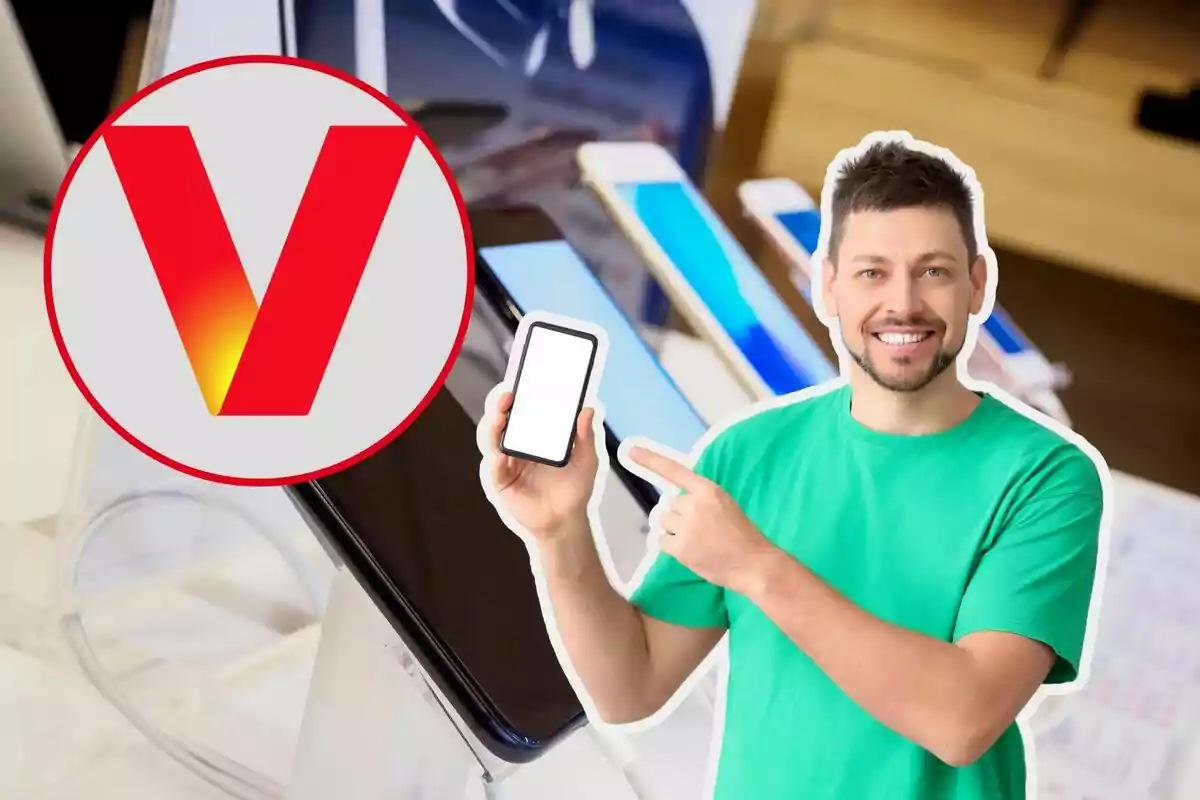 Un hombre sonriente con camiseta verde sostiene y señala un teléfono móvil con pantalla en blanco, junto a un logotipo circular con una letra "V" roja, en un entorno de tienda de tecnología con varios dispositivos en exhibición.