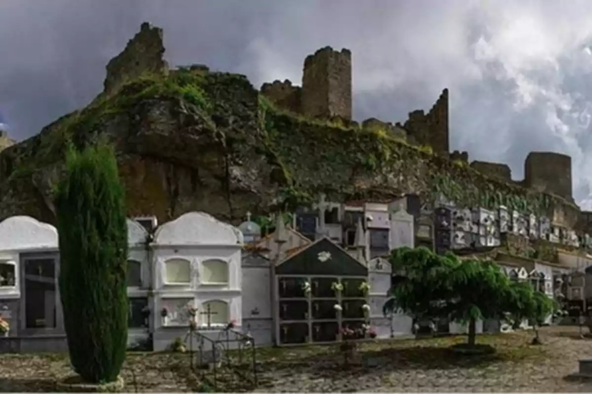 Un cementerio con nichos y tumbas frente a las ruinas de un castillo bajo un cielo nublado.