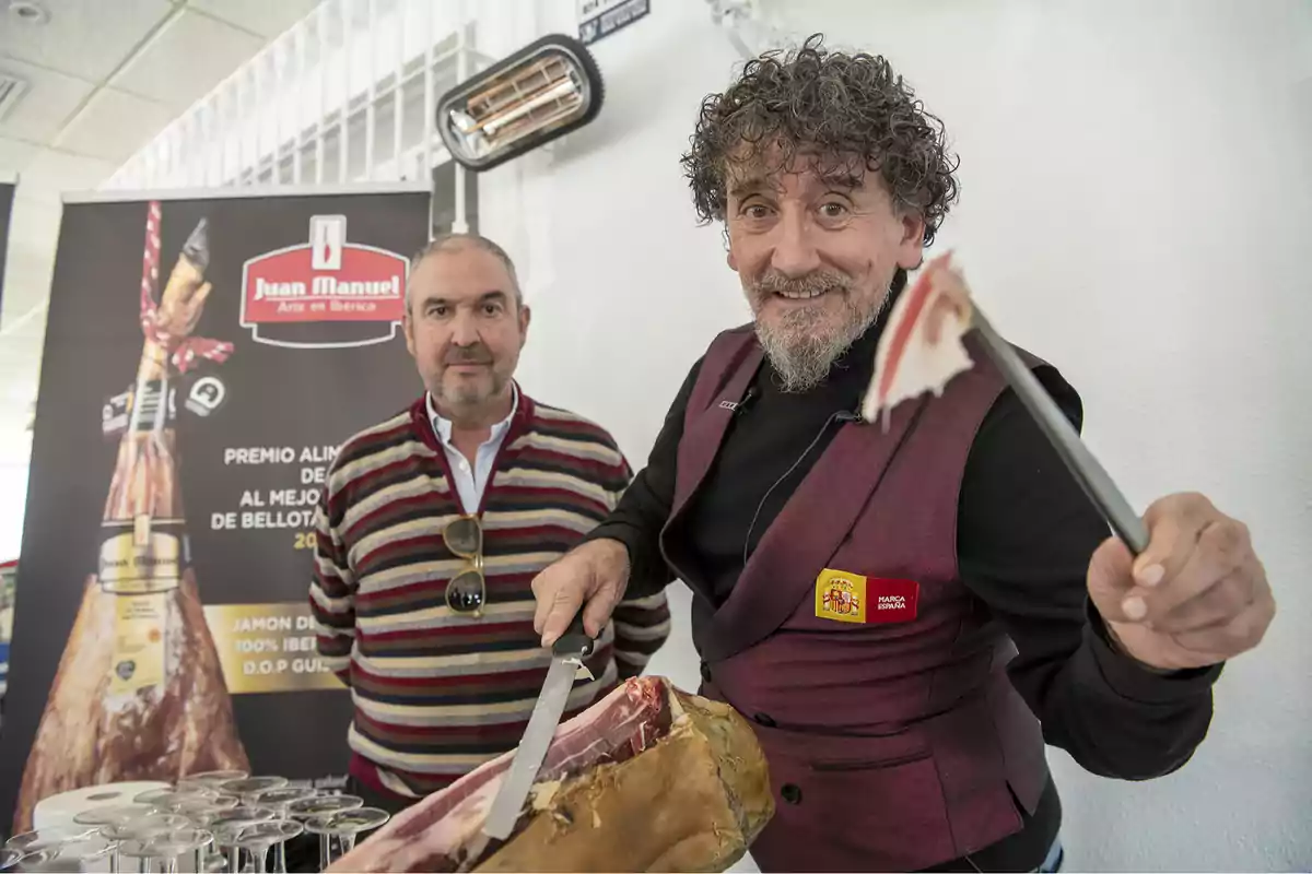 Dos hombres posan junto a un jamón ibérico, uno de ellos sostiene una loncha recién cortada mientras el otro está de pie junto a un cartel promocional.