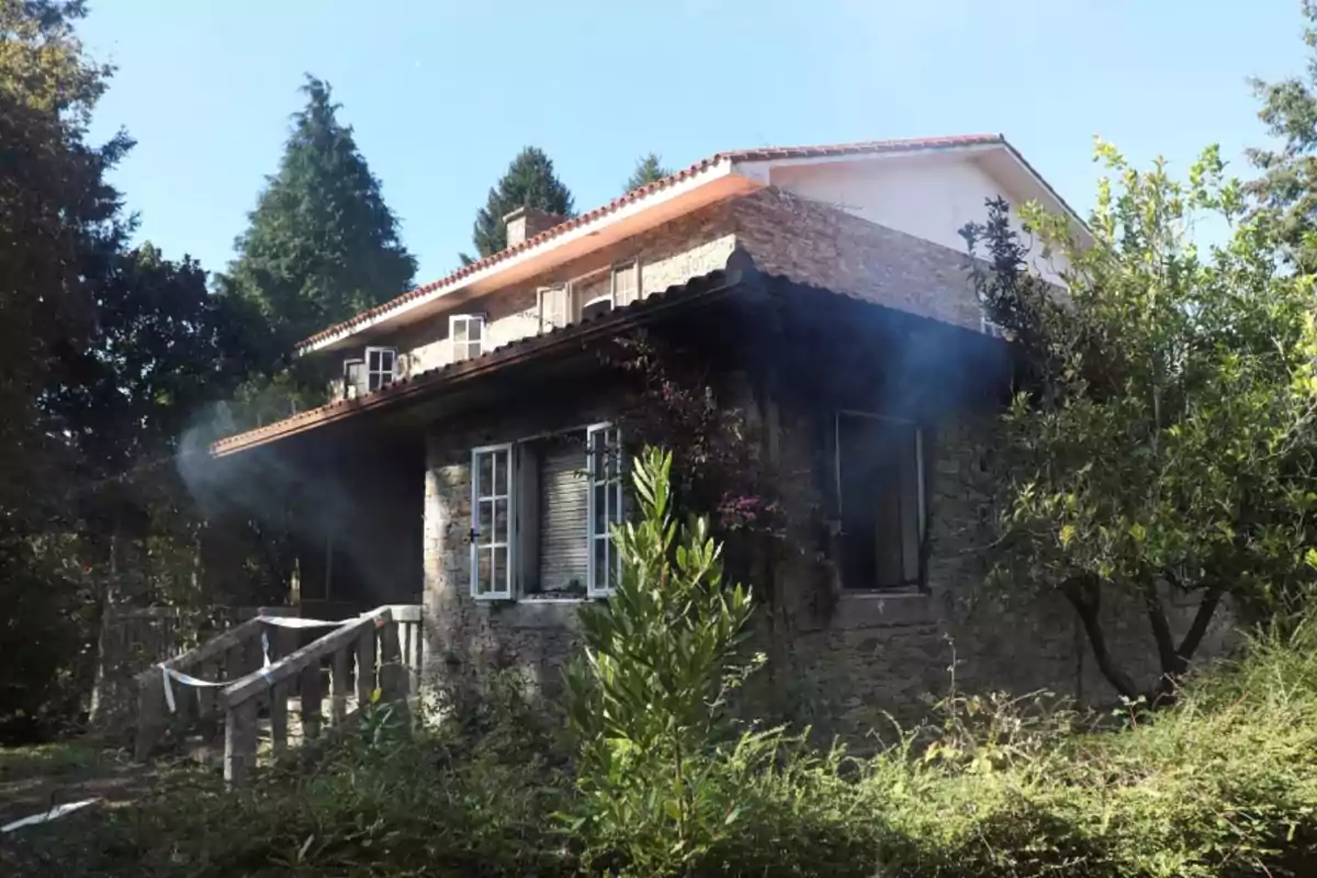 Una casa de piedra con ventanas blancas y un techo de tejas rojas, rodeada de vegetación y árboles, con humo saliendo de su interior.