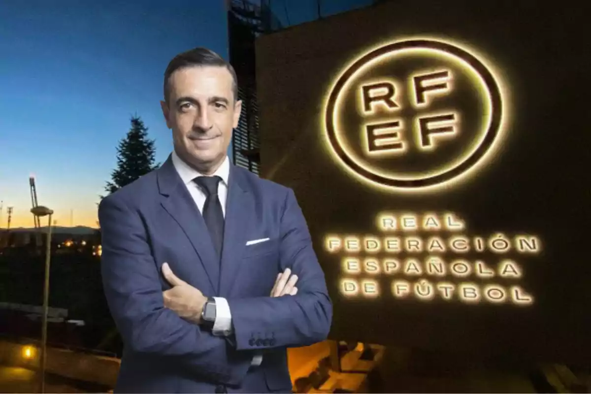 Un hombre con traje y corbata posa frente a un edificio con el logotipo iluminado de la Real Federación Española de Fútbol al atardecer.