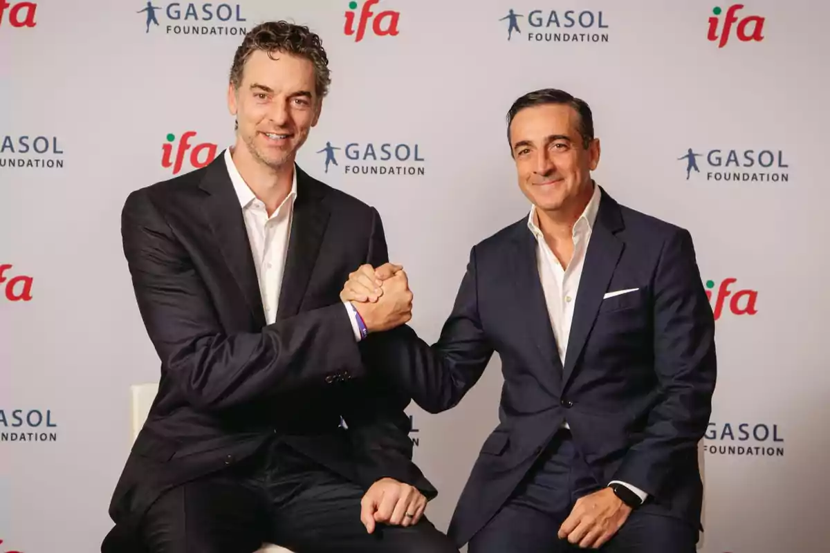 Dos hombres vestidos con trajes oscuros se dan la mano frente a un fondo con los logotipos de Gasol Foundation e IFA.