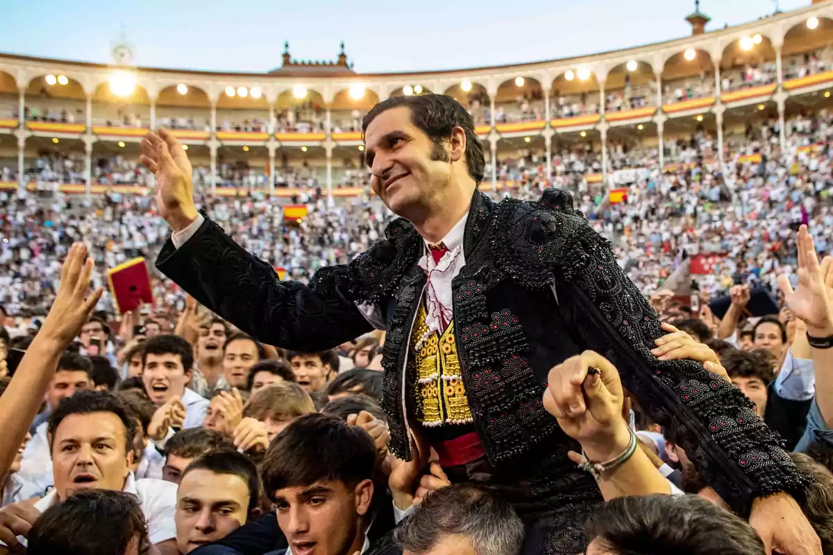 Un torero vestido de traje tradicional es llevado en hombros por una multitud en una plaza de toros llena de espectadores