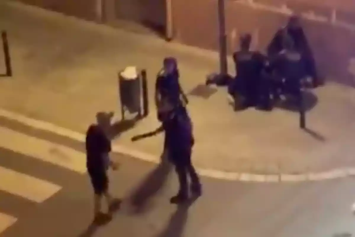 Una persona con uniforme de policía sostiene una porra mientras se enfrenta a otra persona en una calle por la noche, con dos personas sentadas en el fondo.