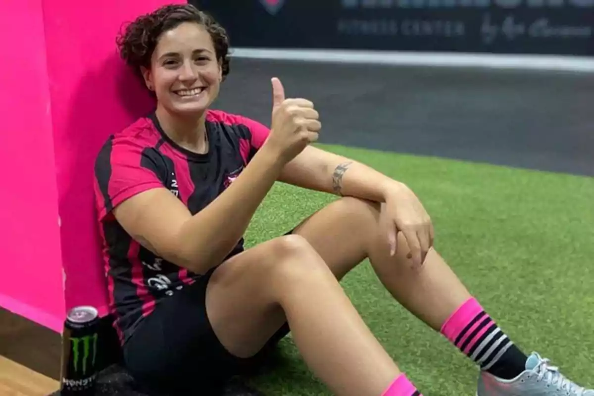 Una persona sentada en el suelo con ropa deportiva, sonriendo y levantando el pulgar, con una lata de bebida energética a su lado.