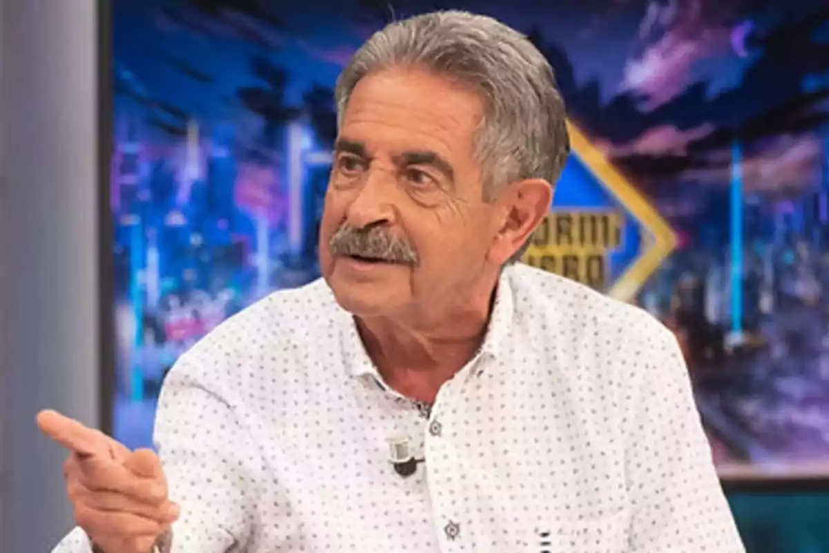 Un hombre de cabello canoso y bigote está hablando en un programa de televisión con un fondo colorido. Un hombre de cabello canoso y bigote está hablando en un programa de televisión con un fondo colorido.