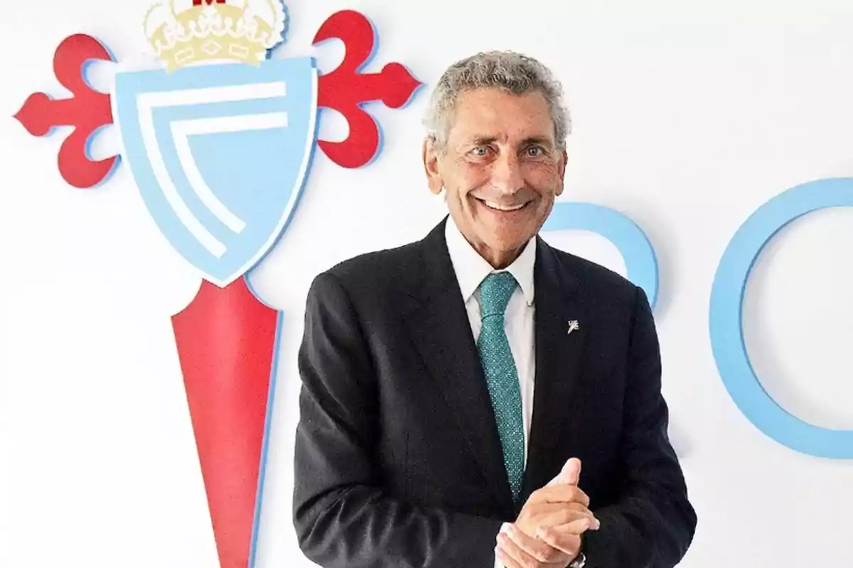 Un hombre sonriente con traje y corbata frente a un escudo de un club deportivo.