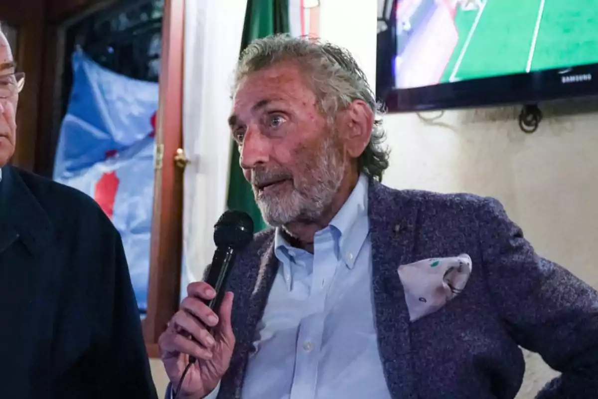 Un hombre mayor con barba y cabello canoso sostiene un micrófono mientras habla en un evento en un interior.