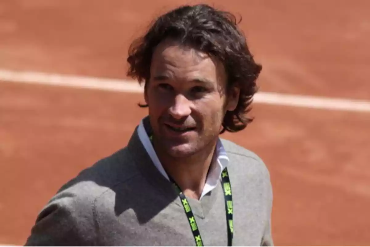 Un hombre con cabello oscuro y rizado lleva un suéter gris y una credencial colgada al cuello mientras está de pie en una cancha de tenis de arcilla. Un hombre con cabello oscuro y rizado lleva un suéter gris y una credencial colgada al cuello mientras está de pie en una cancha de tenis de arcilla.