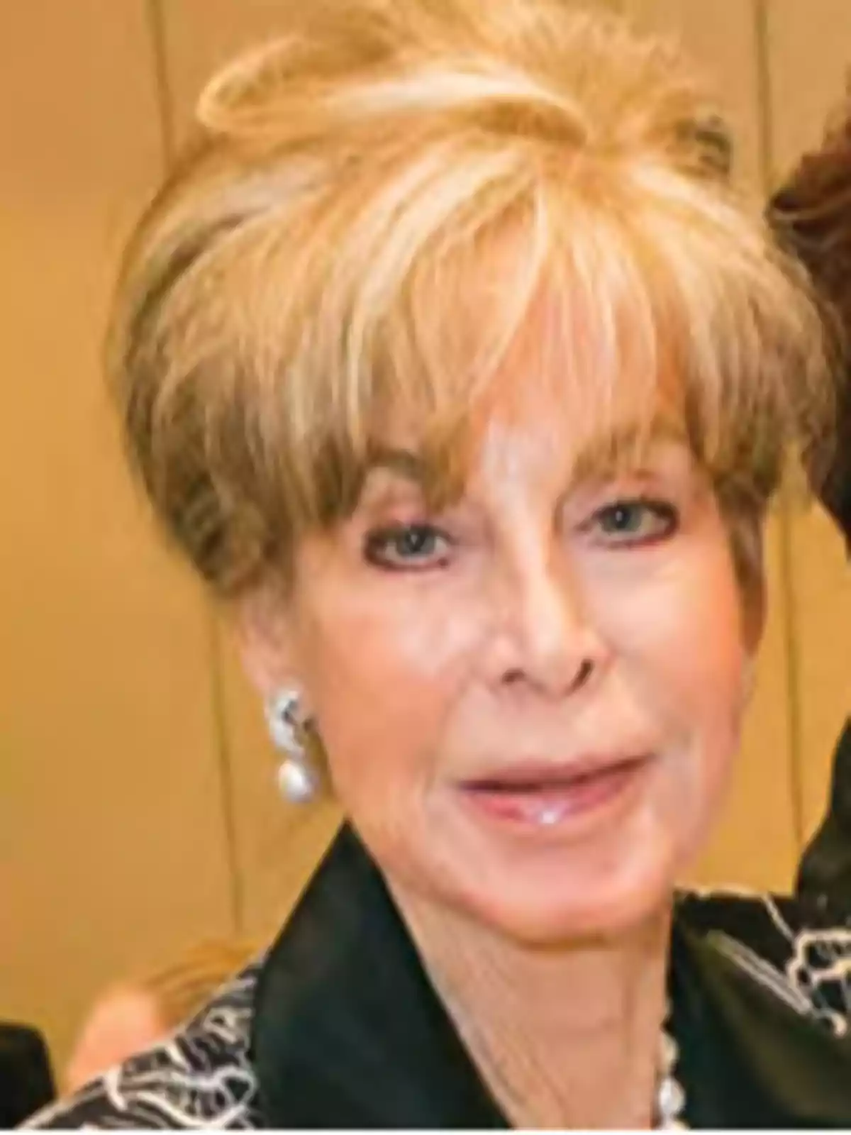 Una persona mayor con cabello rubio y peinado elegante lleva aretes de perlas y una chaqueta oscura. Una persona mayor con cabello rubio y peinado elegante lleva aretes de perlas y una chaqueta oscura.