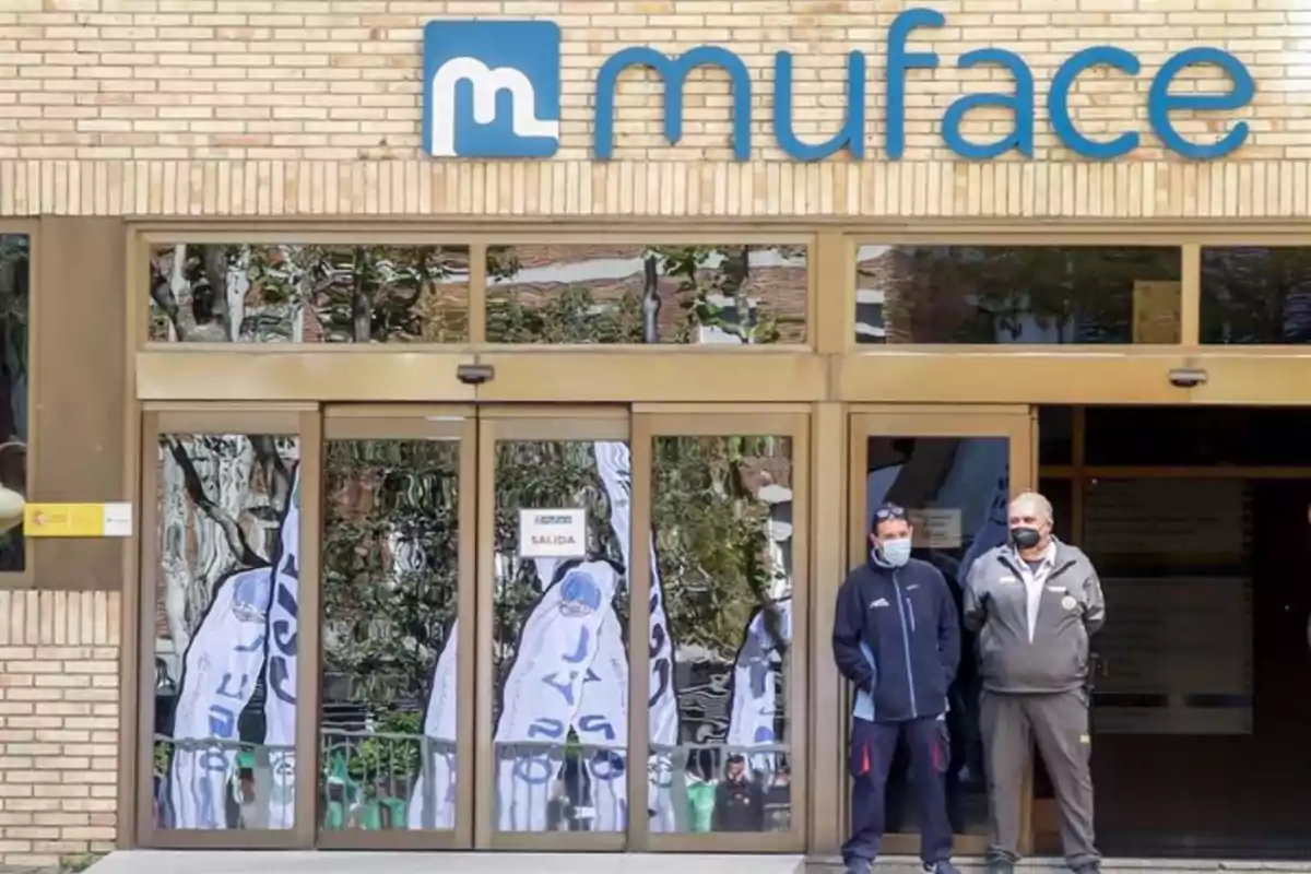 Dos personas con mascarillas están de pie frente a la entrada de un edificio con el letrero "muface".