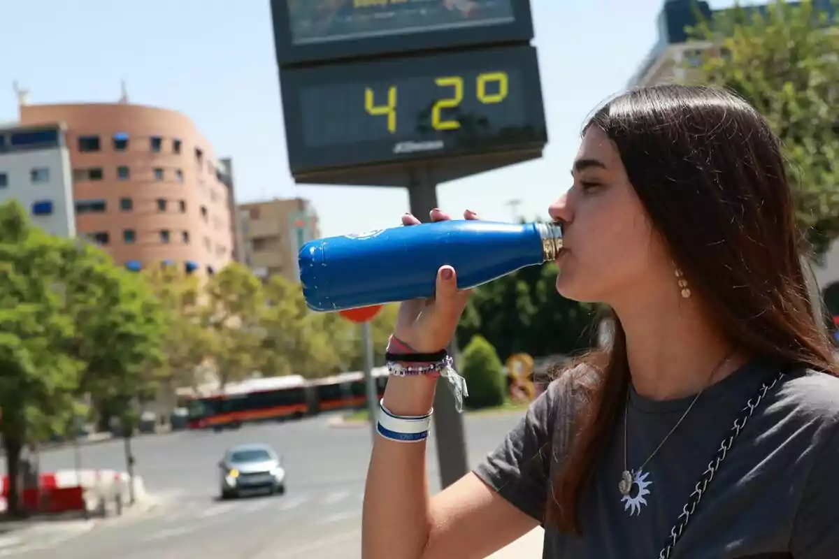 Joven bebiendo agua de una botella azul en la calle durante un día caluroso con un termómetro que marca 42 grados en el fondo. Joven bebiendo agua de una botella azul en la calle durante un día caluroso con un termómetro que marca 42 grados en el fondo.