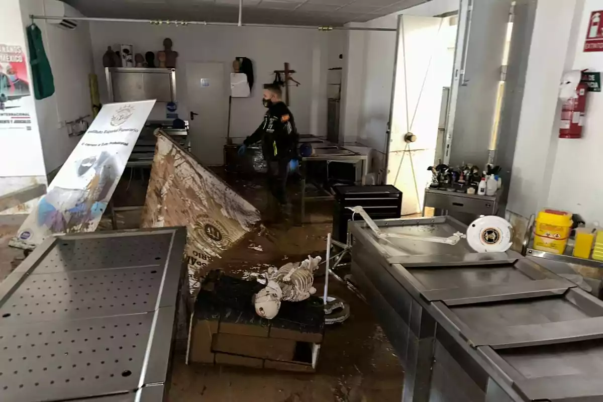 Un hombre con mascarilla y guantes camina por una sala desordenada y llena de barro con muebles metálicos y un esqueleto de práctica en el suelo.