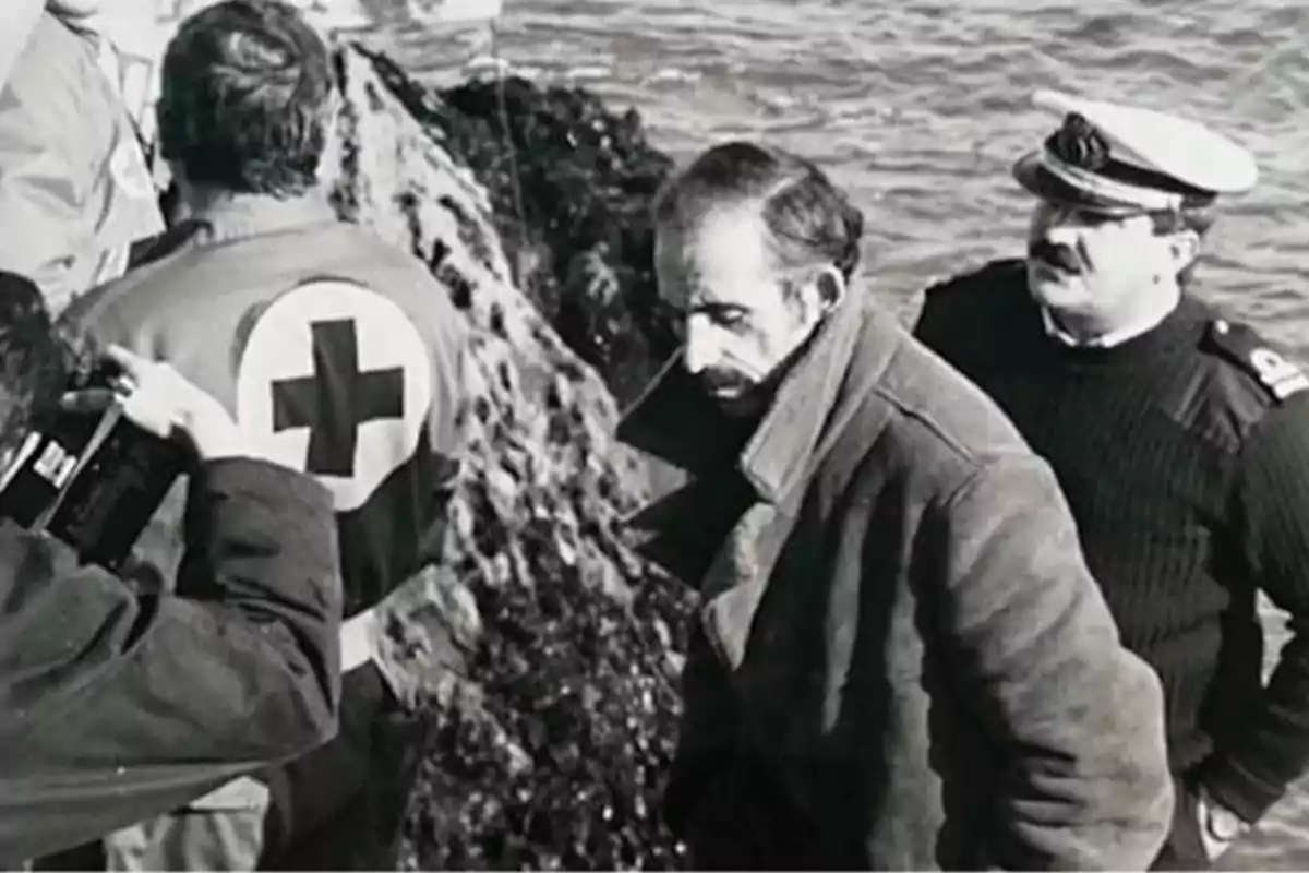 Un grupo de personas, incluyendo un hombre con uniforme de la Cruz Roja y otro con uniforme naval, se encuentra en una zona rocosa junto al mar. Un grupo de personas, incluyendo un hombre con uniforme de la Cruz Roja y otro con uniforme naval, se encuentra en una zona rocosa junto al mar.