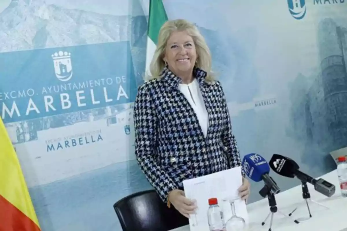 Una mujer de pie en una conferencia de prensa en el Ayuntamiento de Marbella con micrófonos y banderas a su alrededor.