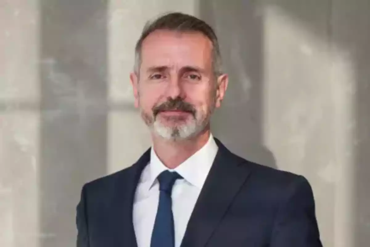 Hombre de mediana edad con barba y cabello corto, vestido con traje oscuro y corbata, posando frente a una pared clara Hombre de mediana edad con barba y cabello corto, vestido con traje oscuro y corbata, posando frente a una pared clara