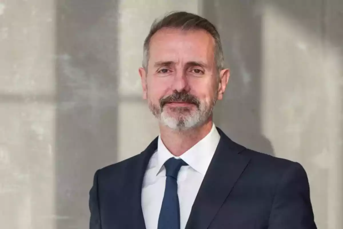 Un hombre de mediana edad con barba y cabello canoso lleva un traje oscuro y corbata, posando frente a un fondo de pared gris.