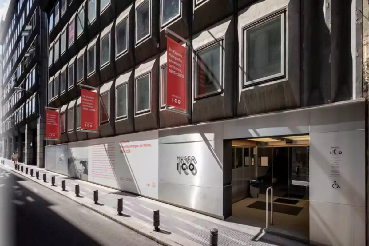 Fachada del Museo ICO en una calle estrecha con carteles rojos de exposición y entrada accesible Fachada del Museo ICO en una calle estrecha con carteles rojos de exposición y entrada accesible