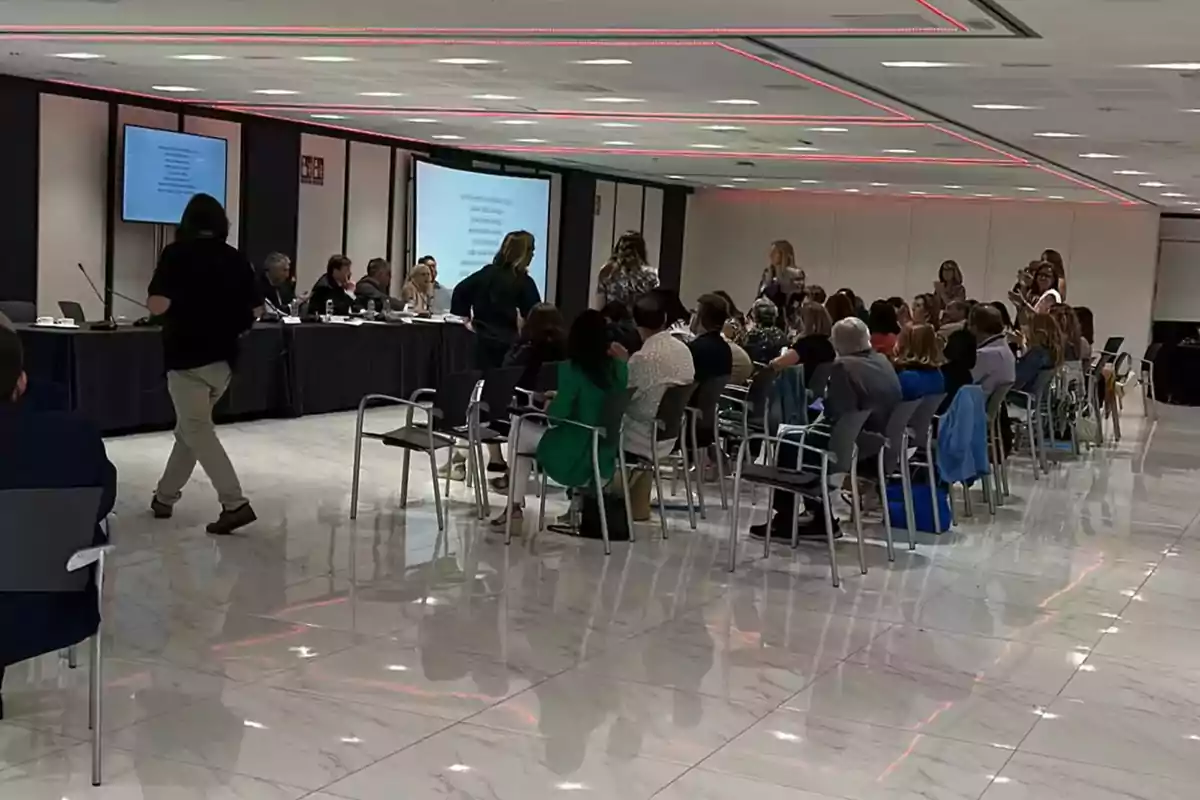 Una reunión o conferencia en una sala moderna con personas sentadas en sillas y varias personas de pie, con una mesa larga al frente donde se encuentran varios oradores y pantallas proyectando información en la pared.