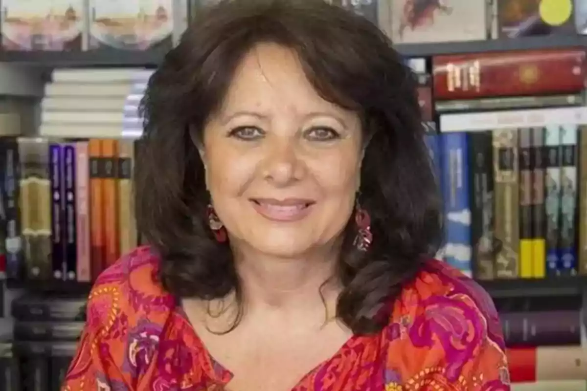 Una mujer sonriente con cabello oscuro y una blusa colorida, sentada frente a una estantería llena de libros.