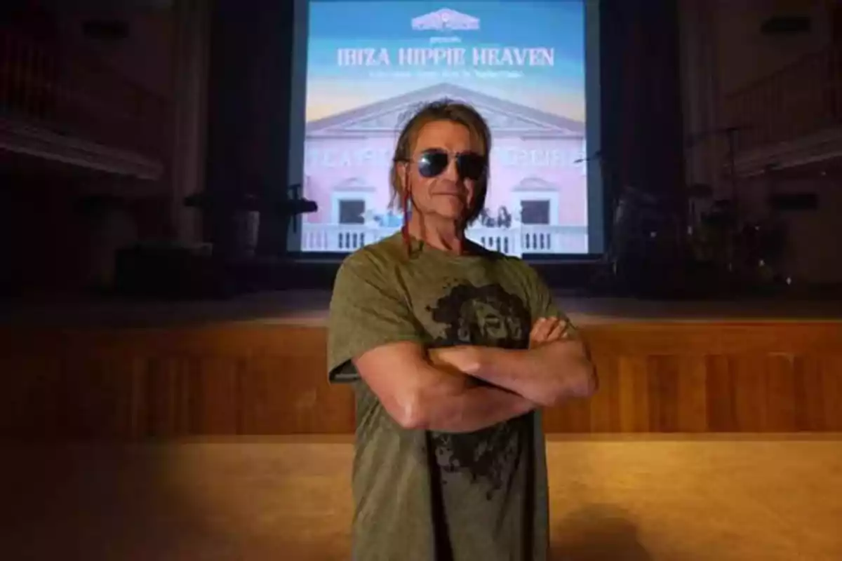 Persona con gafas de sol y camiseta verde posando con los brazos cruzados frente a una pantalla que muestra el texto "Ibiza Hippie Heaven".