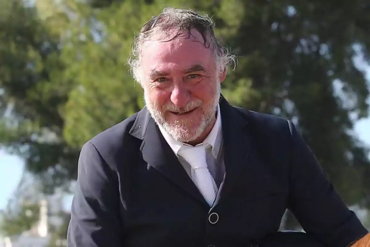 Hombre mayor con barba y cabello canoso, vestido con traje oscuro y corbata blanca, sonriendo al aire libre con árboles de fondo.