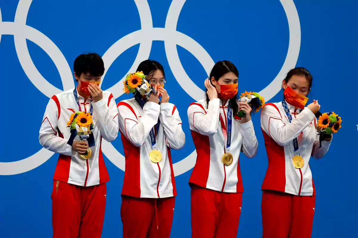 Cuatro atletas con medallas de oro y ramos de flores en el podio olímpico, vistiendo uniformes rojos y blancos, con un fondo de los anillos olímpicos.