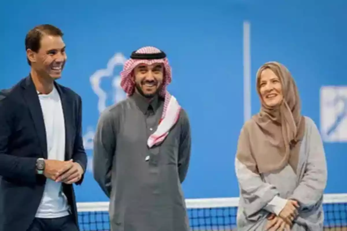 Tres personas sonrientes posan frente a una cancha de tenis con un fondo azul.