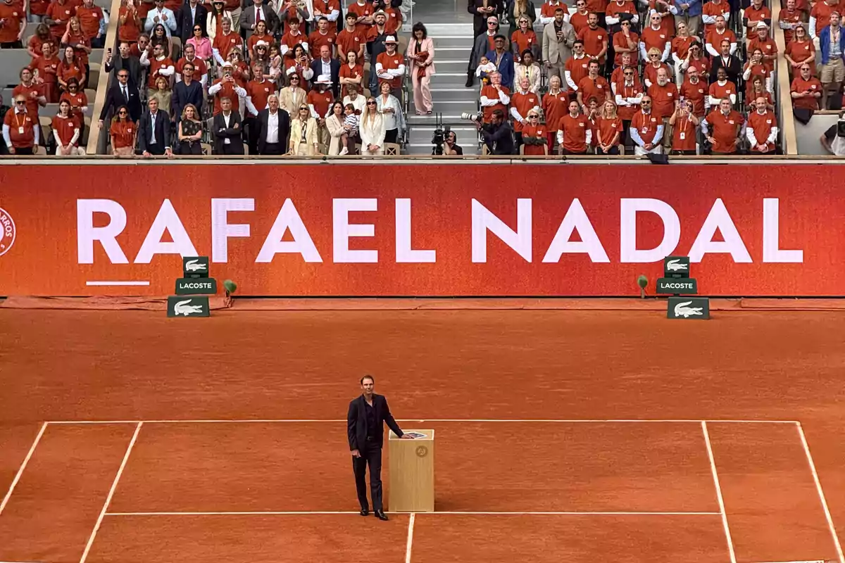 Un hombre de pie junto a un podio en una cancha de tenis con una pantalla gigante detrás que muestra un nombre, rodeado de una multitud de personas en las gradas. Un hombre de pie junto a un podio en una cancha de tenis con una pantalla gigante detrás que muestra un nombre, rodeado de una multitud de personas en las gradas.