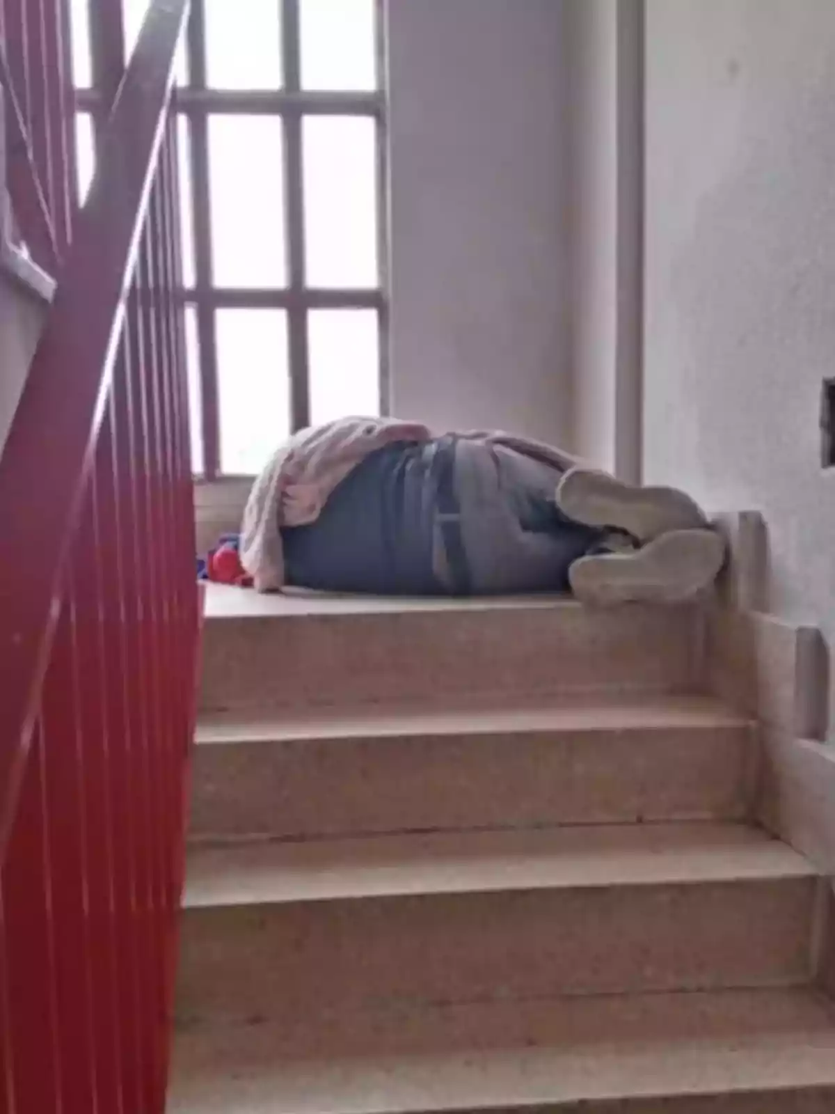 Persona acostada en las escaleras junto a una ventana con barandilla roja.