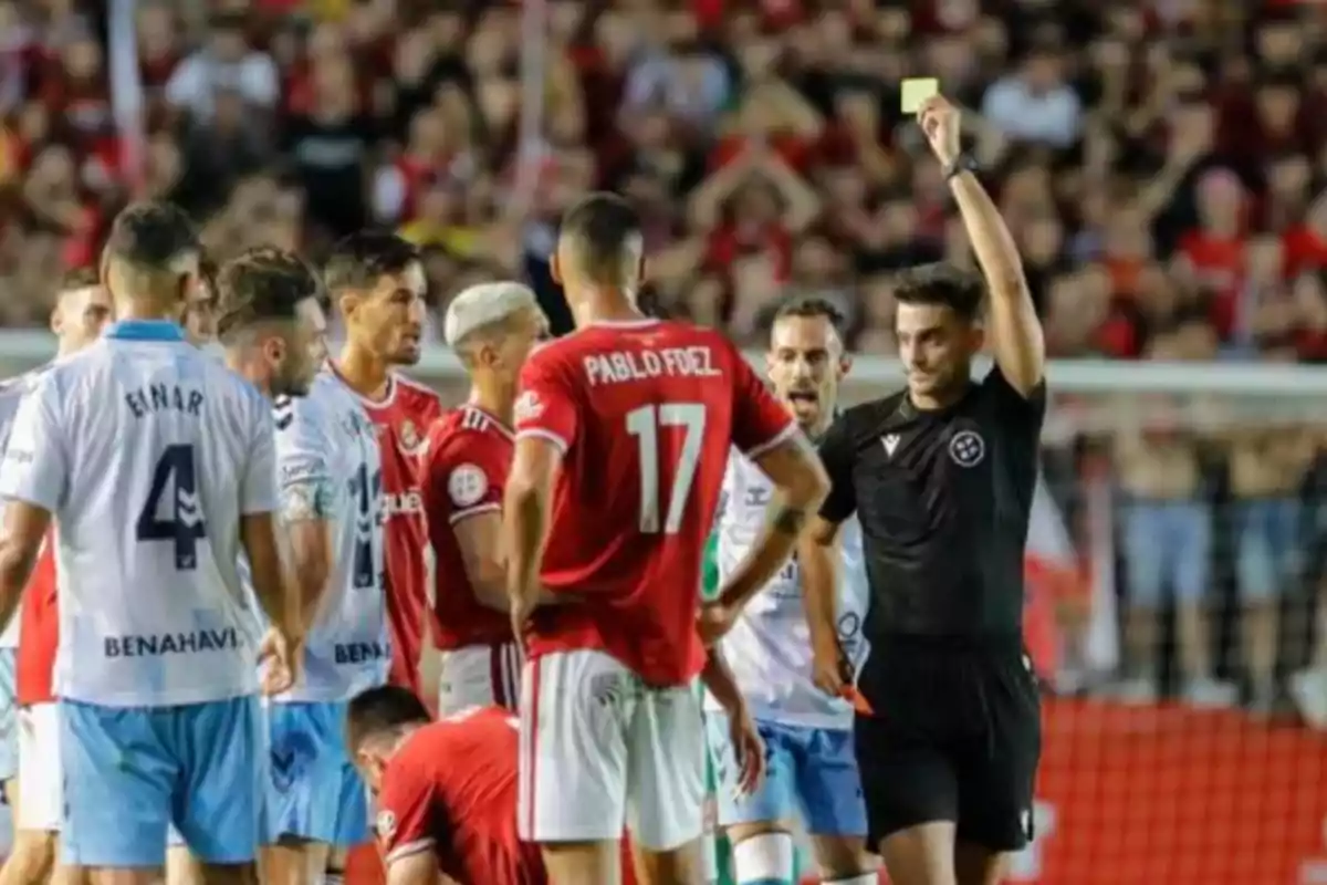 Un árbitro muestra una tarjeta amarilla a un jugador durante un partido de fútbol mientras otros jugadores observan la situación.