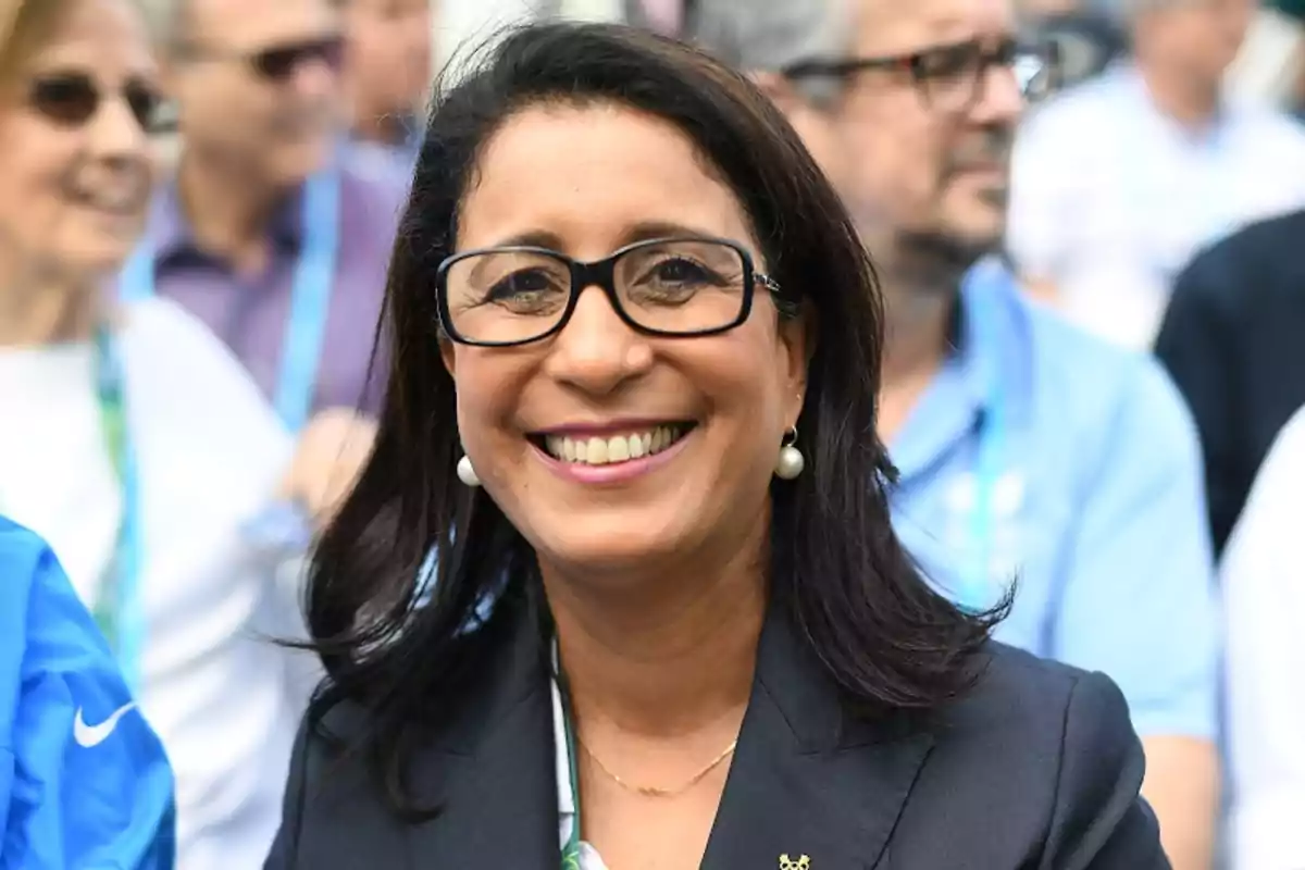 Una mujer sonriente con gafas y aretes de perla, vestida con un blazer oscuro, rodeada de personas en un evento al aire libre.