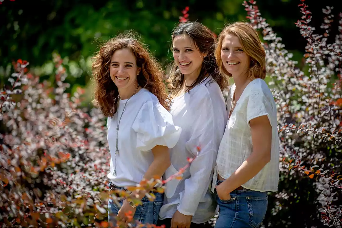 Tres mujeres sonrientes posando al aire libre entre arbustos y vegetación.