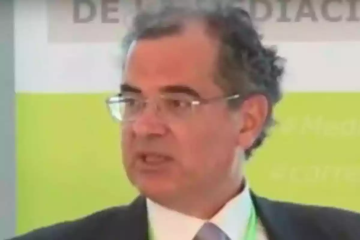 Hombre con gafas y cabello canoso hablando en un evento con fondo verde y blanco.