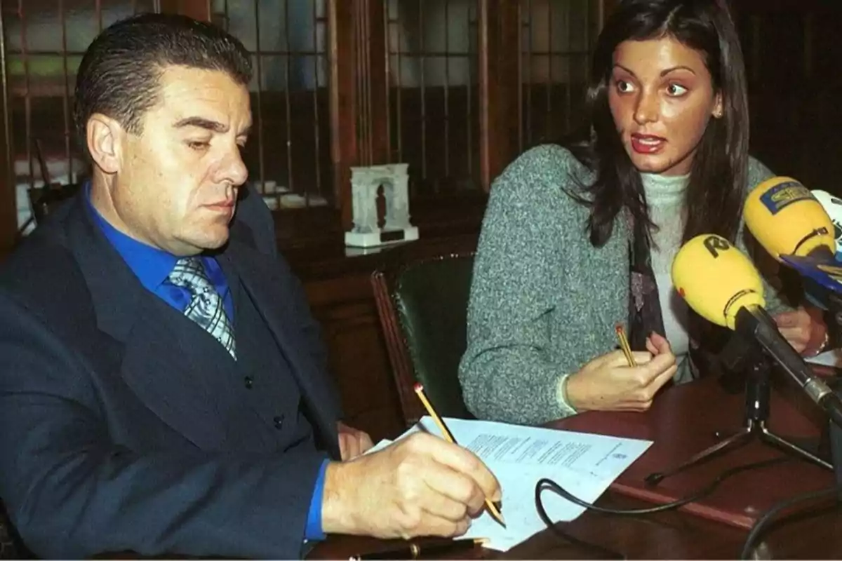 Dos personas sentadas en una mesa, una de ellas firmando un documento mientras la otra habla frente a varios micrófonos.