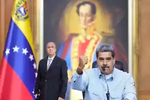 Un hombre de pie con traje oscuro y corbata, junto a otro hombre sentado con camisa azul claro, levantando el puño, con una bandera de Venezuela y un retrato en el fondo.